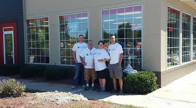 About Us photo with 3 individuals standing next to each other in front of large windows and a small green hedge below the window.
